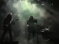 Un groupe de metal sur scène, avec un guitariste à gauche, un chanteur au centre, et un batteur à droite. La scène est enfumée, éclairée par des projecteurs.
