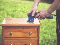 Un homme utilise une ponceuse pour le décapage mécanique d'une commode en bois, préparant le meuble pour une nouvelle finition.