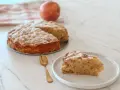 Une part de **crépiau aux pommes** saupoudrée de sucre glace, à côté d'un gâteau entier et d'une pomme. Une cuillère dorée est posée sur l'assiette.