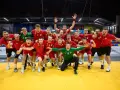Drużyna piłkarzy ręcznych w czerwonych strojach świętuje zwycięstwo. Wśród nich Krzysztof Zimny, bramkarz w zielonym stroju, klęczy z wyciągniętymi rękami.