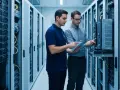 Two IT professionals review network infrastructure technologies on a tablet amidst rows of servers in a data center.
