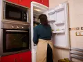 Kobieta w fartuchu zagląda do lodówki. Obok stoi piekarnik, a nad nim wbudowana mikrofalówka. Czy mikrofalówka może stać na lodówce? To pytanie, na które odpowiedź znajdziesz w instrukcji urządzenia.