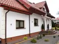 Biały dom z brązowymi cokołami, dachówką i rynnami. Przed domem schody, ozdobne krzewy i ścieżka z kostki brukowej.
