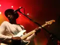 Marcus Miller w czarnym kapeluszu gra na białej gitarze basowej na tle czerwonych świateł scenicznych.