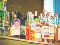 Rząd butelek z alkoholem, w tym gin i inne trunki, gotowe do serwowania. Wśród nich widać butelkę z etykietą "No Mistake Old Tom Gin", a także inne, które mogą zawierać ajerkoniak.