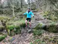 Kvinna springer i skogen, fokuserad på att springa marathon. Mossbeklädda stenar och träd skapar en utmanande terräng.