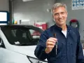 Uśmiechnięty mechanik w granatowym kombinezonie podaje kluczyki do białego samochodu. W tle warsztat samochodowy, gdzie wszystko jest gotowe, by sprawdzić, co to homologacja.