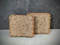 Zwei Scheiben rustikales **Sauerteig Kastenbrot** mit Körnern und einer luftigen Krume vor dunklem Hintergrund.