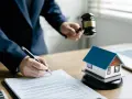 Licytacja elektroniczna nieruchomości: młotek sędziego i model domu na biurku, gdzie podpisuje się dokumenty.
