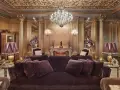 Salón lujoso con sofás de terciopelo morado, lámparas doradas y un candelabro de cristal. El techo y las paredes doradas evocan el estilo barroco español.