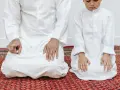 Ojciec i syn w białych strojach klęczą na dywanie, wykonując codzienną modlitwę poranną.