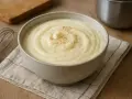 Bol de béchamel onctueuse saupoudré de poivre, avec un fouet et une planche en bois en arrière-plan.