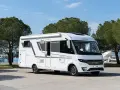 En vit Adria Sonic husbil, den lyxigaste husbilen, parkerad vid en marina med båtar och träd i bakgrunden.