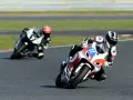 Dwóch motocyklistów w pełnym rynsztunku ściga się na torze. Jeden z nich, z numerem 11, pochyla się nisko w zakręcie, demonstrując emocje sportu motocyklowego.