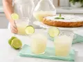 Dos vasos de granizado de limón refrescante, adornados con rodajas de lima, junto a limones frescos y un pastel.