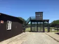 Brama i wieża strażnicza na terenie Muzeum Stutthof. Drewniane budynki i brukowany chodnik.