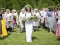 Grupa kobiet wiankach na głowach i tradycyjnych strojach idzie przez łąkę. Obraz nawiązuje do święta zielone świątki.