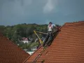 Robotnik w kasku i szelkach bezpieczeństwa na dachu, pracujący przy dachówce.