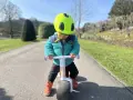 A child in a bright green helmet and teal jacket rides a woom wow balance bike on a sunny path.