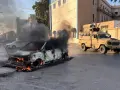 Płonący samochód i pojazd opancerzony na ulicy. Obraz sugeruje, czy Libia jest bezpieczna, pokazując chaos i obecność wojska.