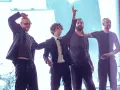 Matty Healy i zespół The 1975 na scenie. Muzycy pozdrawiają publiczność, jeden z nich unosi rękę w geście pokoju.