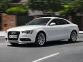 Białe Audi A5 Coupe pędzi po drodze, jego sylwetka jest dynamiczna i elegancka.
