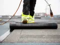 Pracownik z palnikiem montuje papę podkładową na dachu.