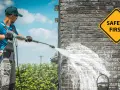 Man demonstrates how to use a pressure washer on a brick wall, with a "Safety First" sign visible.