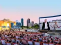 Ein lauer Sommerabend im Freiluftkino Charlottenburg. Menschen sitzen in Liegestühlen und schauen auf eine Leinwand, auf der ein Film mit einem älteren Mann und einer jungen Frau läuft. Im Hintergrund die Berliner Skyline.