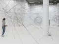 Une femme admire une installation de Tomas Saraceno, un réseau complexe de fils noirs tendus dans un espace blanc.