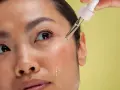 Kropla serum z pipetki spada na policzek kobiety. To może być retinol, który co to jest i jak działa, warto sprawdzić.