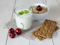 Un verre de yaourt lait végétal, garni de menthe, avec des cerises et des crackers sur une table en bois.