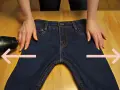 Manos sujetan pantalones vaqueros oscuros sobre mesa de madera. Secador de pelo apunta a la tela. Aprende como estirar pantalones vaqueros.