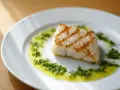 Un delicioso bacalao a la plancha, con marcas doradas y servido sobre un plato blanco con aceite de oliva y hierbas frescas picadas.