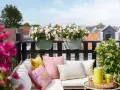Ein gemütlicher Balkon mit bunten Kissen, gelber Kanne und blühenden sommerblumen.