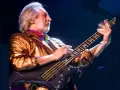 Ein Mann mit grauem Haar und Bart spielt Bassgitarre. Er trägt eine glänzende goldene Jacke und bunte Krawatte. Er ist der beste Bassist der Welt.