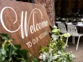 Drewniana tablica "Welcome to our wedding" ozdobiona zielenią. W tle widać przygotowane stoły i krzesła na uroczystość.