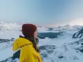 Kvinna i gul jacka och röd mössa njuter av utsikten över snöklädda berg och hav. Perfekt för sportlov i Norge.