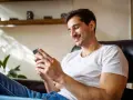 Uśmiechnięty mężczyzna w białej koszulce siedzi w fotelu i patrzy na telefon.