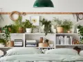 Grüne Pflanzen im Schlafzimmer schaffen eine beruhigende Atmosphäre. Auf weißen Regalen stehen diverse Pflanzen in Körben und Töpfen, daneben Bücher und Deko.