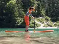 Kobieta w pomarańczowych spodenkach i sportowej bluzce uprawia pływanie na supie po spokojnym jeziorze. W tle zielone drzewa i jasna plaża.