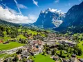 A scenic view of Grindelwald, Switzerland, nestled in a valley with lush green hills and a majestic snow-capped mountain. A train travels through the village.