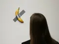 Dojrzały banan przyklejony do ściany szarą taśmą, widziany zza ramienia osoby z długimi, ciemnymi włosami.
