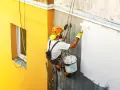 Malowanie elewacji cena za m2: alpinista maluje ścianę budynku na biało, wisząc na linach.