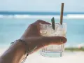 Orzeźwiający drink na plaży, z lodem i plasterkiem limonki, w dłoni z bransoletką. W tle błękitne morze i fale.
