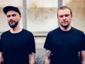 Dwóch mężczyzn w czarnych koszulkach. Jeden w czapce z daszkiem, drugi z zaczesanymi włosami. Ich tatuaże, jak tworzywo sztuczne za mało czasu tekst, opowiadają historie.