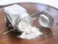 Un barattolo di vetro rovesciato su un tavolo di legno, con polvere bianca che fuoriesce, forse ingredienti per colla vinilica fatta in casa.