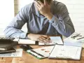 Zestaw dokumentów, kalkulator i telefon leżą na biurku. Mężczyzna z ręką na czole, przytłoczony problemami finansowymi, przegląda pismo windykacyjne.