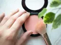 Une main applique un pinceau sur un blush poudre, révélant une douce teinte couleur blush.