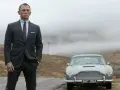 Uomo con impeccabile 007 outfit, completo gessato e cravatta, posa accanto a una Aston Martin d'epoca in un paesaggio nebbioso.